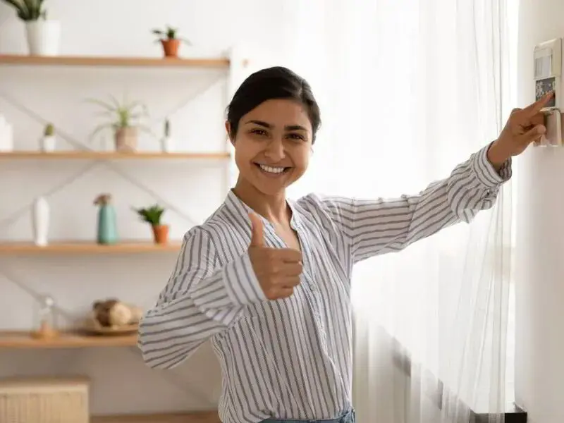 A smiling woman adjusts a thermostat, giving a thumbs up gesture